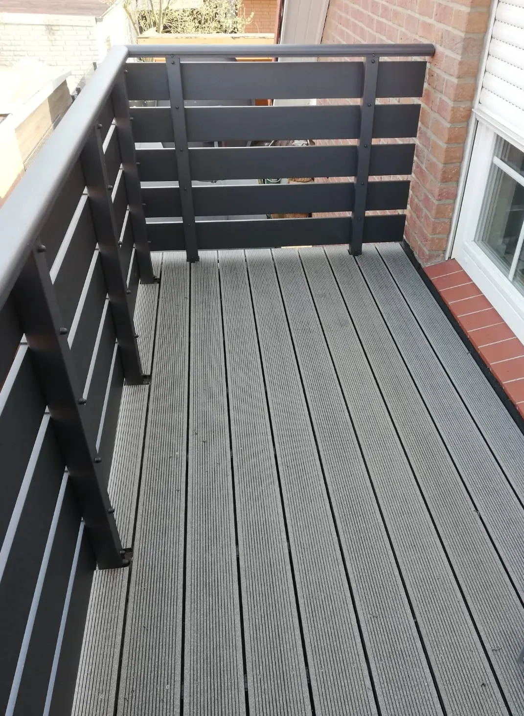 Fertige WPC-Dielen in Grau mit anthrazitfarbenem Geländer auf einem Balkon an einer Backsteinfassade